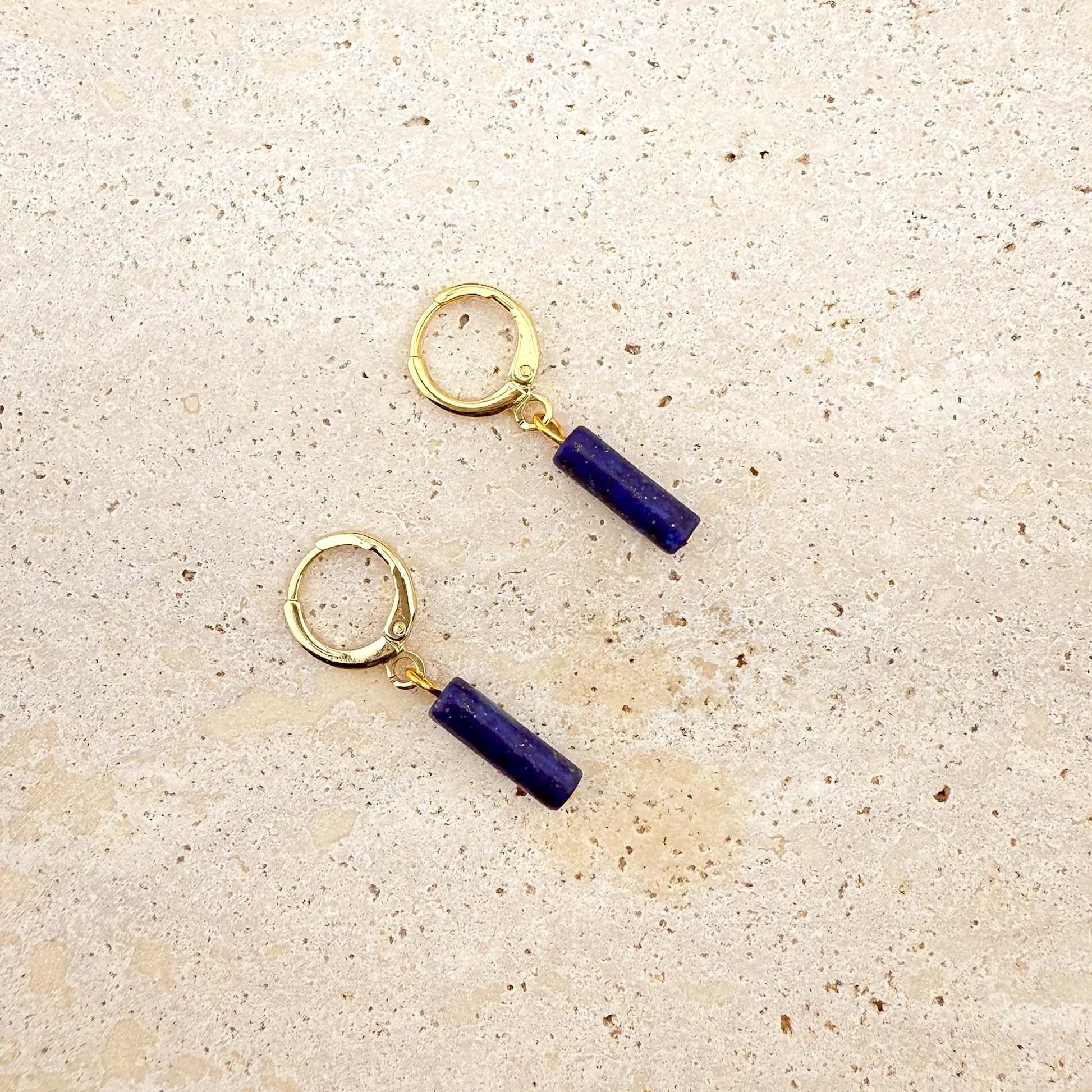 A pair of gold Aking Sinta huggie hoop earrings featuring deep blue lapis lazuli column charms. The natural gemstone cylinders showcase a rich ultramarine hue with subtle gold pyrite flecks, suspended from polished gold-toned hoops on a light-colored natural stone surface.