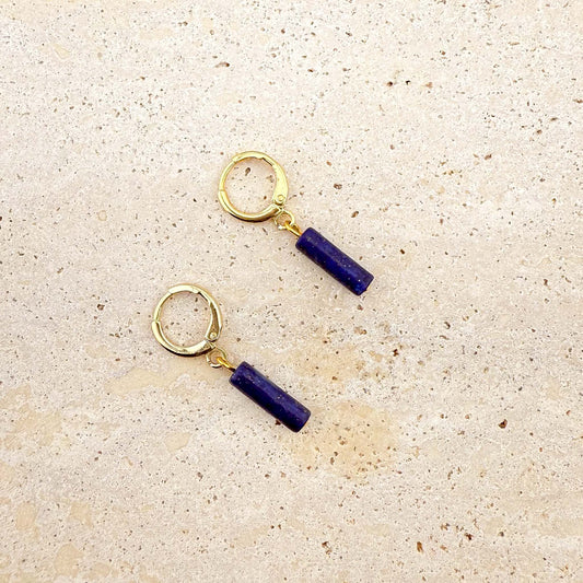 A pair of gold Aking Sinta huggie hoop earrings featuring deep blue lapis lazuli column charms. The natural gemstone cylinders showcase a rich ultramarine hue with subtle gold pyrite flecks, suspended from polished gold-toned hoops on a light-colored natural stone surface.