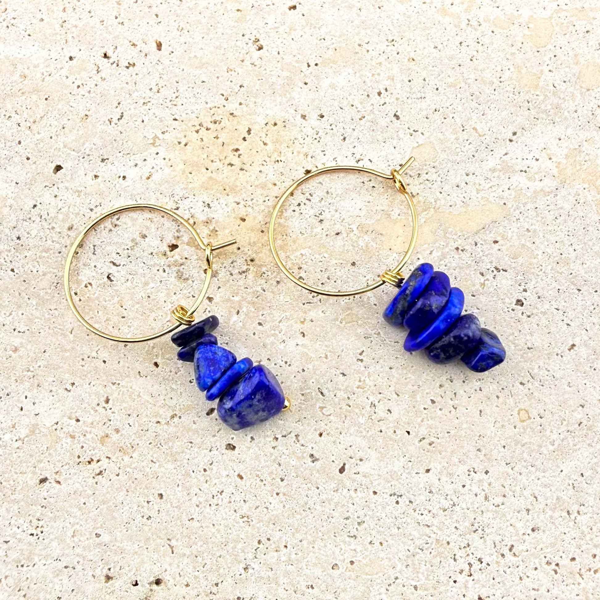 Pair of Aking Sinta gold huggie earrings with raw natural Lapis Lazuli nugget charms on a white marble background.