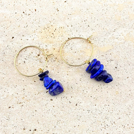 Pair of Aking Sinta gold huggie earrings with raw natural Lapis Lazuli nugget charms on a white marble background.