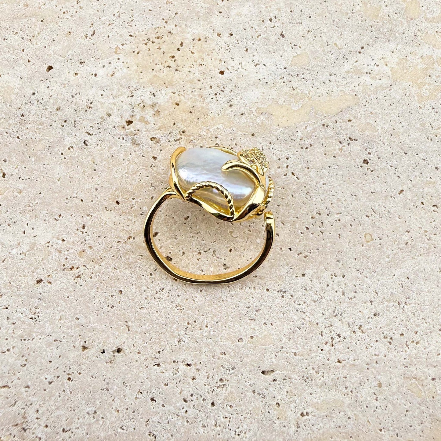 A side-angle view of an adjustable Aking Sinta gold ring featuring a lustrous, organic Keshi pearl. The ring band and the intricate gold wire setting, embellished with a sparkling pave-set gold leaf, are clearly visible against a light, textured natural stone background