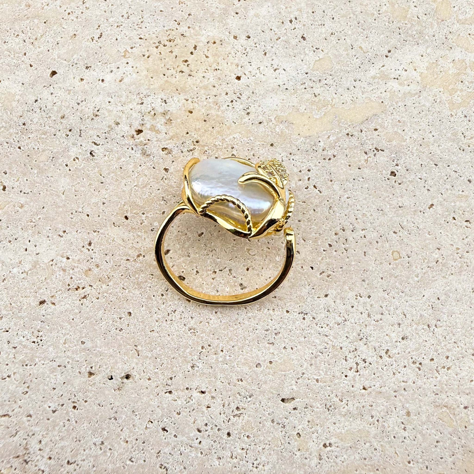A side-angle view of an adjustable Aking Sinta gold ring featuring a lustrous, organic Keshi pearl. The ring band and the intricate gold wire setting, embellished with a sparkling pave-set gold leaf, are clearly visible against a light, textured natural stone background