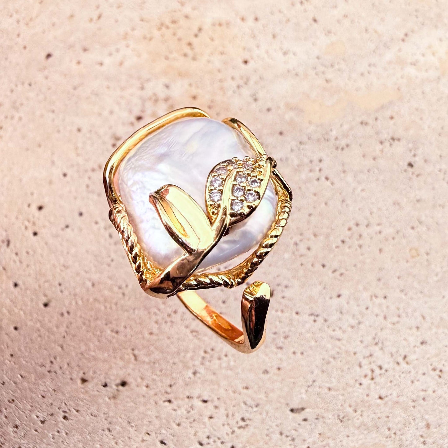 A top-down view of an adjustable Aking Sinta gold ring, showcasing a large, organic-shaped Keshi pearl framed in twisted gold wire. The piece is elegantly accented with a delicate, sparkling pave-set gold leaf, presented against a light, textured natural stone surface