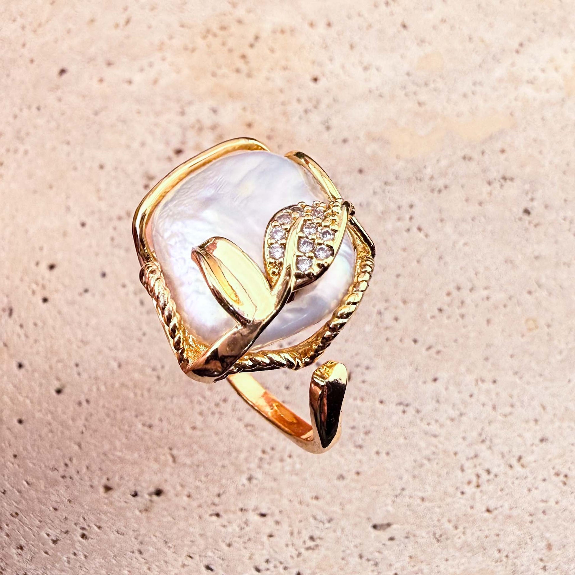 A top-down view of an adjustable Aking Sinta gold ring, showcasing a large, organic-shaped Keshi pearl framed in twisted gold wire. The piece is elegantly accented with a delicate, sparkling pave-set gold leaf, presented against a light, textured natural stone surface