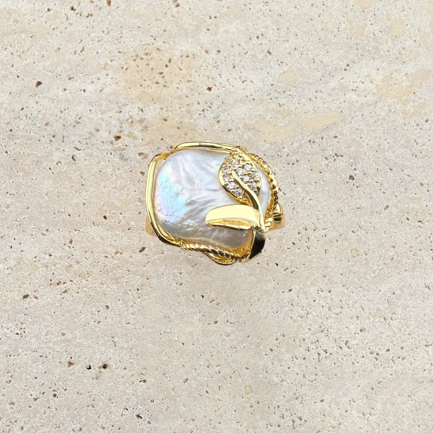 A detailed close-up of an adjustable Aking Sinta ring featuring a large, iridescent, organic-shaped Keshi pearl. The pearl is framed by polished gold wire and embellished with a delicate, sparkling pave-set gold leaf accent. The ring is showcased on a light, textured natural stone surface
