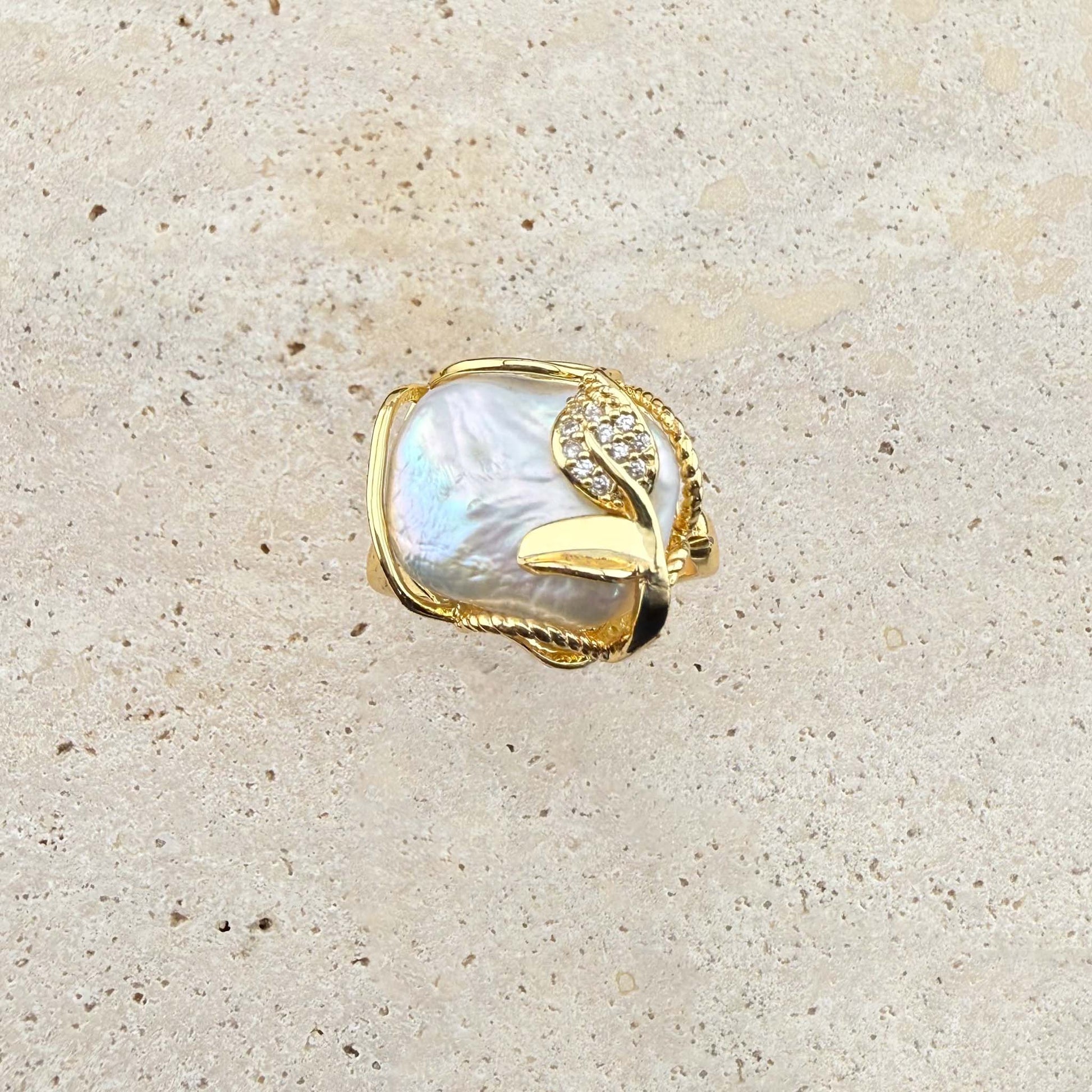 A detailed close-up of an adjustable Aking Sinta ring featuring a large, iridescent, organic-shaped Keshi pearl. The pearl is framed by polished gold wire and embellished with a delicate, sparkling pave-set gold leaf accent. The ring is showcased on a light, textured natural stone surface