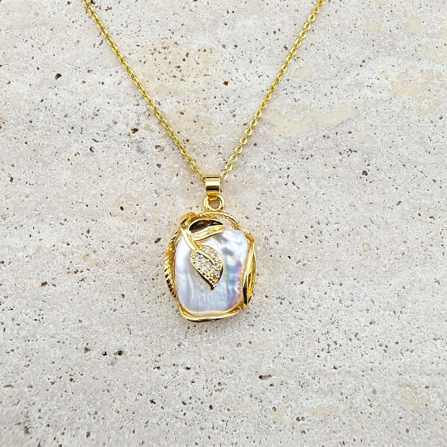 A gold Aking Sinta necklace featuring a luminous, organic-shaped Keshi pearl pendant delicately wrapped in gold wire and accented with a small, shimmering pave-set gold leaf charm. The pendant hangs from a fine gold chain, presented against a light, textured natural stone surface