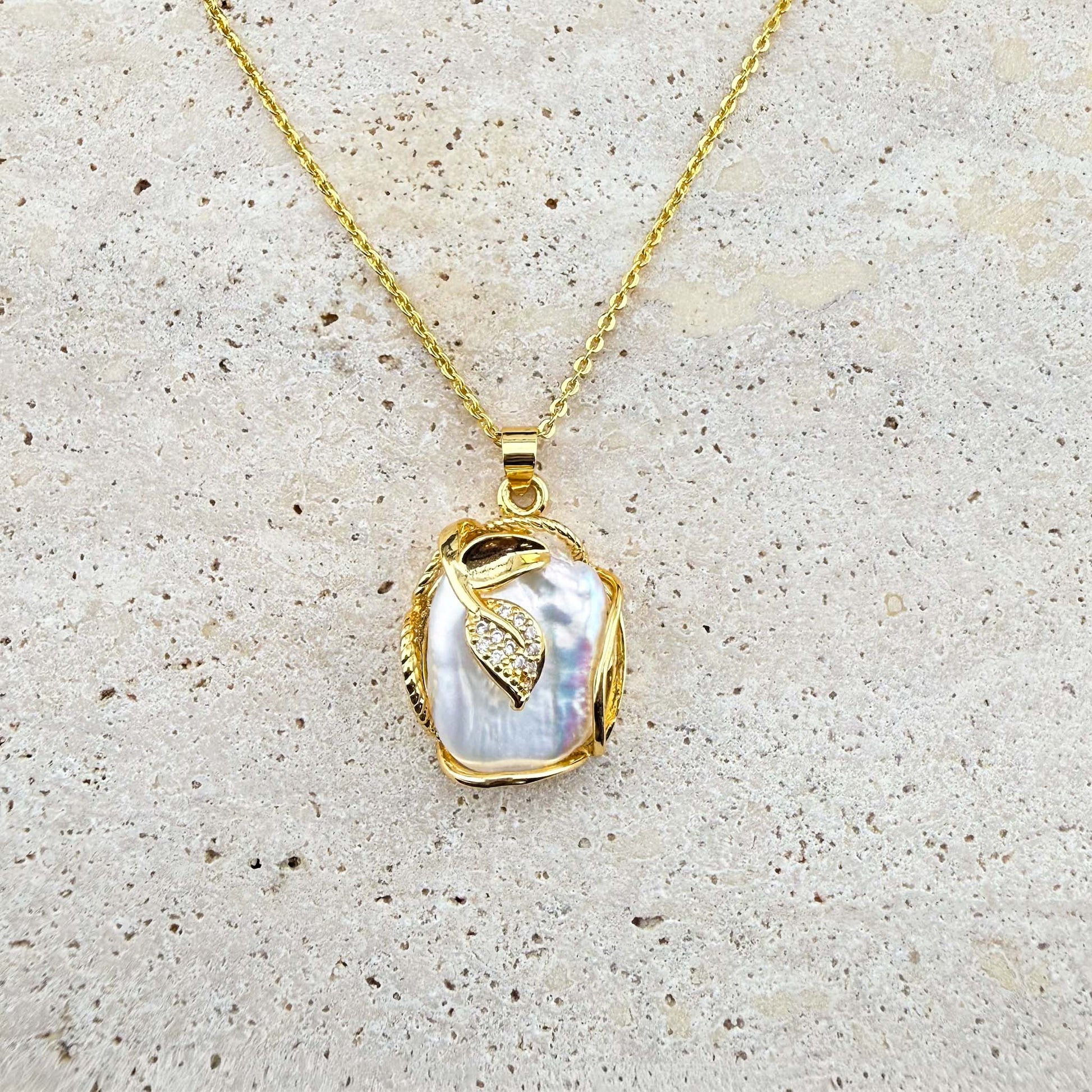 A gold Aking Sinta necklace featuring a luminous, organic-shaped Keshi pearl pendant delicately wrapped in gold wire and accented with a small, shimmering pave-set gold leaf charm. The pendant hangs from a fine gold chain, presented against a light, textured natural stone surface