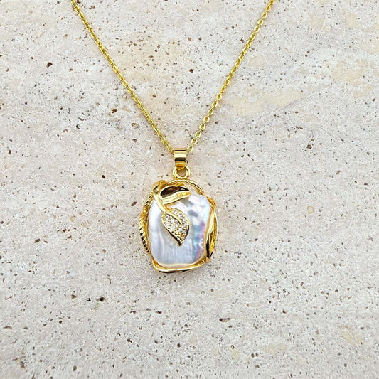 A gold Aking Sinta necklace featuring a luminous, organic-shaped Keshi pearl pendant delicately wrapped in gold wire and accented with a small, shimmering pave-set gold leaf charm. The pendant hangs from a fine gold chain, presented against a light, textured natural stone surface