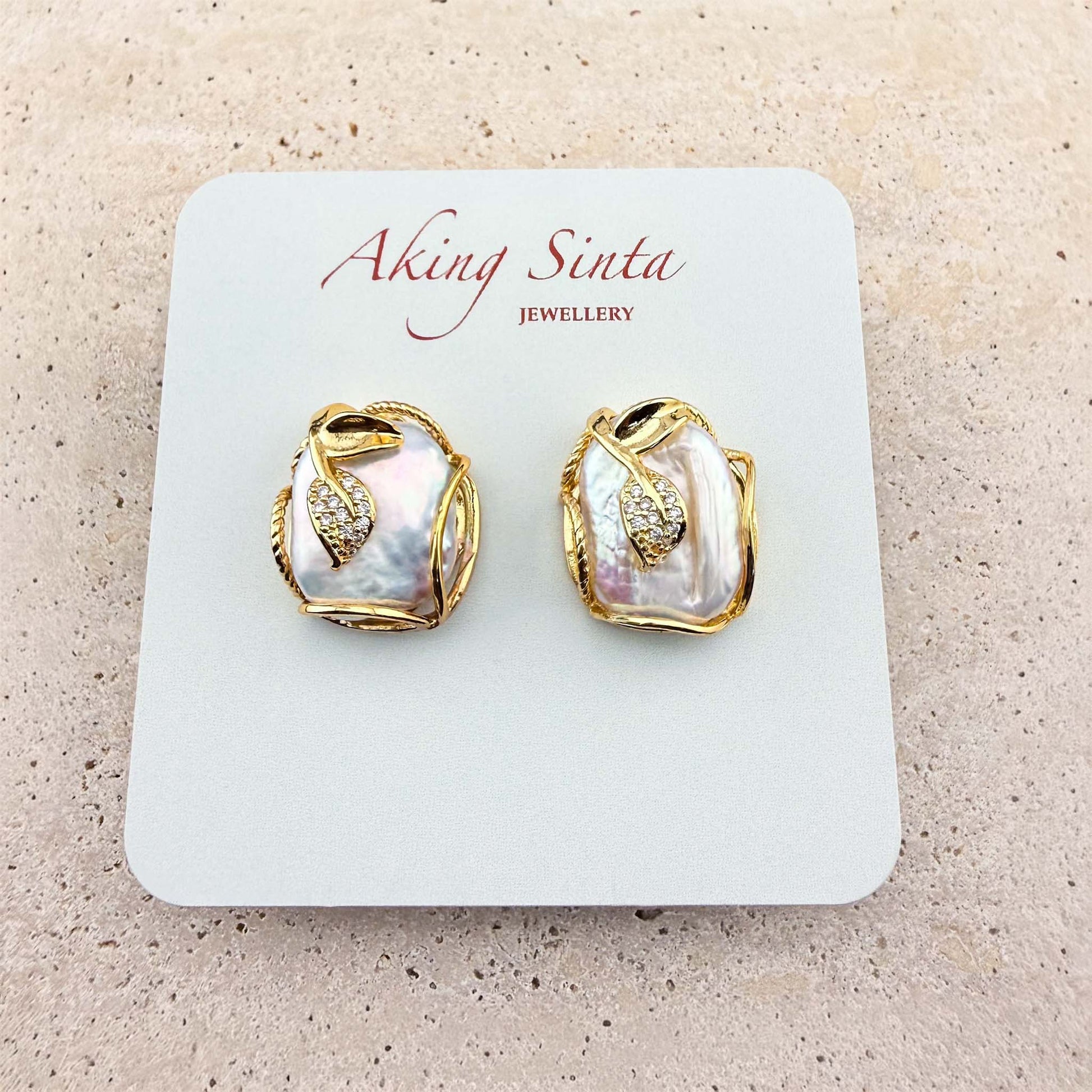 A pair of Aking Sinta stud earrings presented on a branded display card. The earrings feature organic-shaped, iridescent Keshi pearls delicately encased in gold wire, each accented with a small, sparkling pave-set gold leaf charm. The earrings are displayed on a light, textured natural stone surface