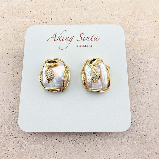 A pair of Aking Sinta stud earrings presented on a branded display card. The earrings feature organic-shaped, iridescent Keshi pearls delicately encased in gold wire, each accented with a small, sparkling pave-set gold leaf charm. The earrings are displayed on a light, textured natural stone surface