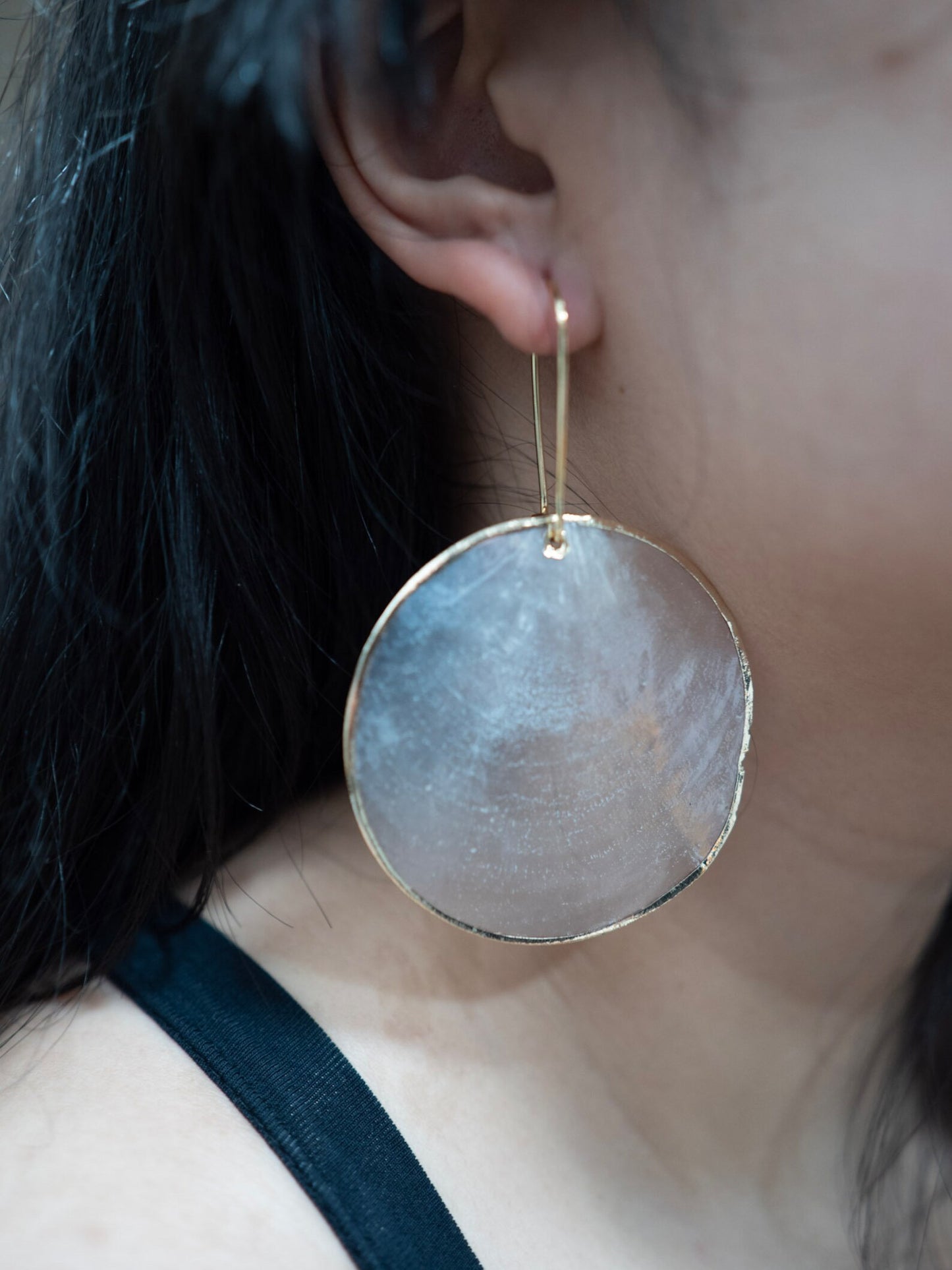 A close-up profile of a woman’s left ear wearing an Aking Sinta Statement Gold Lined Capiz Shell earring; the large, round iridescent white disk is framed in polished gold and hangs elegantly, catching the light to show its pearlescent texture.