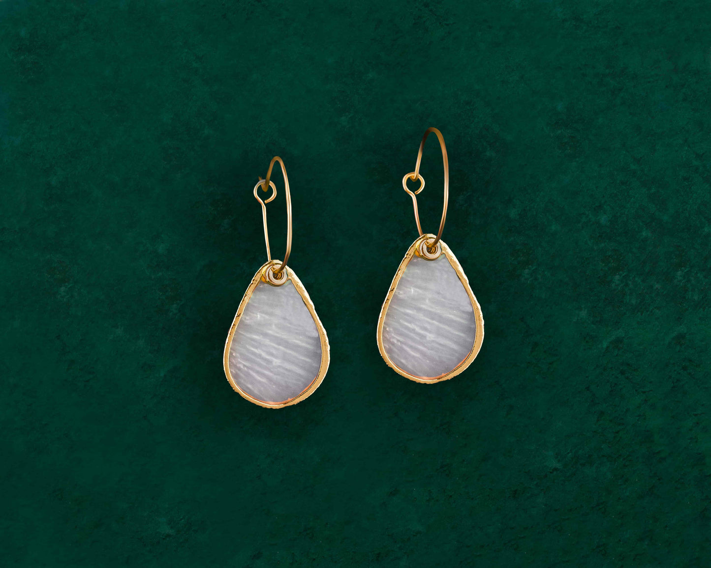 A pair of Aking Sinta Gold Lined Capiz Shell Earrings in a teardrop shape, displayed on a dark green background; featuring elongated, iridescent white Capiz shell drops encased in a polished gold metal trim and hanging from gold ear wires.