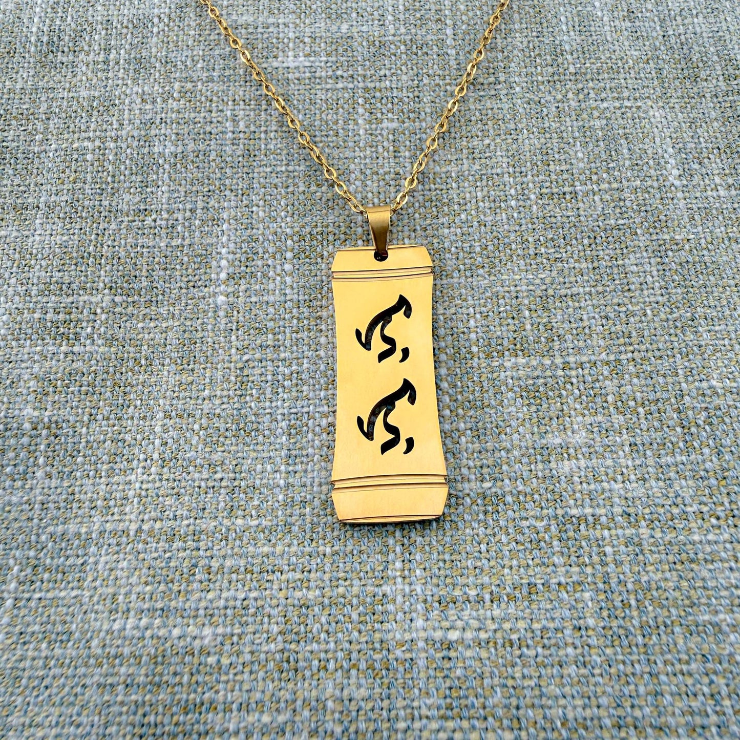 An Aking Sinta gold vertical pendant necklace centered on a textured blue-gray woven linen backdrop. The matte gold rectangular pendant features a stylized bamboo frame outline with the word "Lolo" (Grandfather) intricately cut out in traditional script-like Baybayin characters, hanging from a fine gold link chain