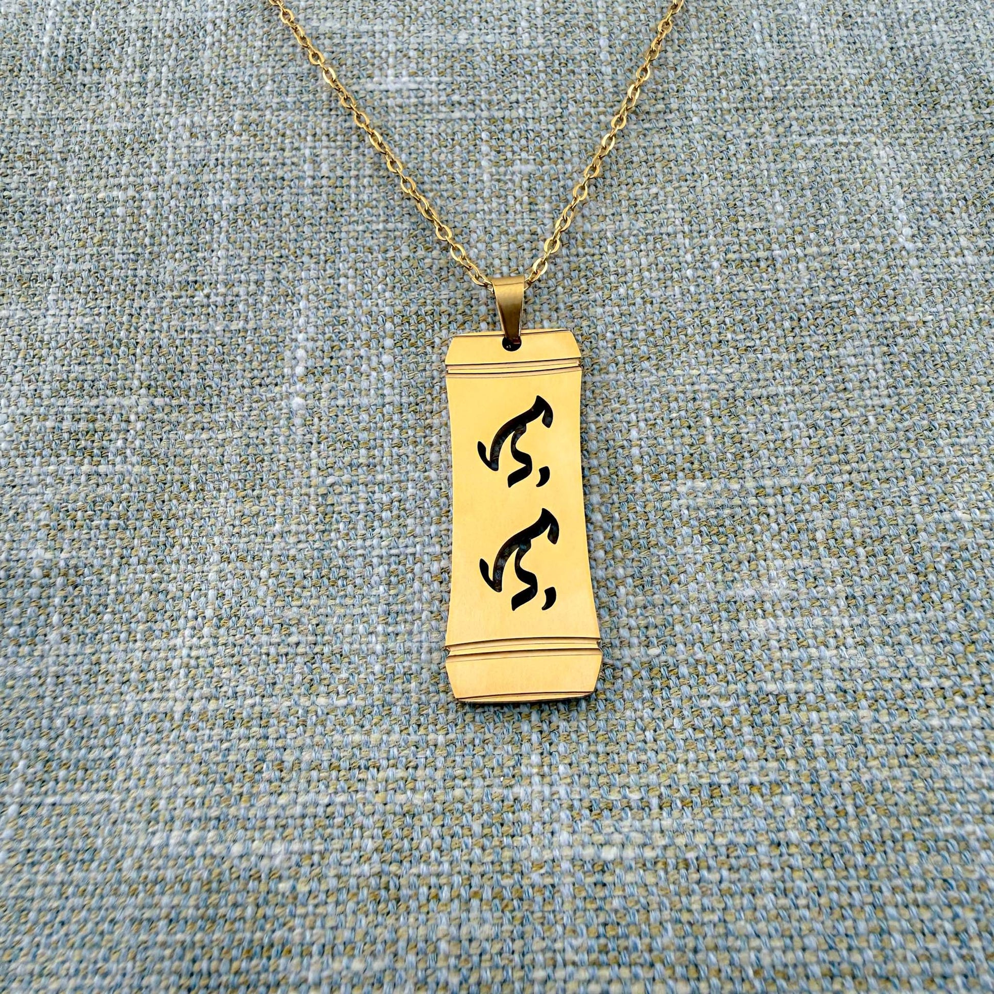 An Aking Sinta gold vertical pendant necklace centered on a textured blue-gray woven linen backdrop. The matte gold rectangular pendant features a stylized bamboo frame outline with the word "Lolo" (Grandfather) intricately cut out in traditional script-like Baybayin characters, hanging from a fine gold link chain