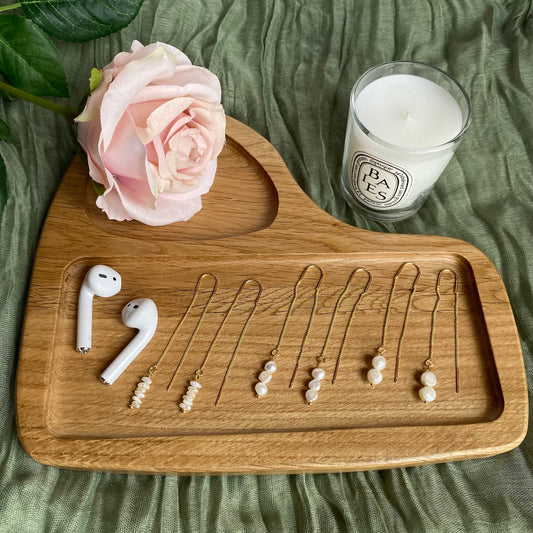 A curated flat-lay photograph featuring three pairs of delicate Aking Sinta gold chain threader earrings, each accented with dainty natural pearls of varying sizes. The earrings are arranged on a minimalist wooden tray alongside a pair of white wireless earbuds, a pale pink faux rose, and a scented candle, all set against a soft, textured olive green fabric background