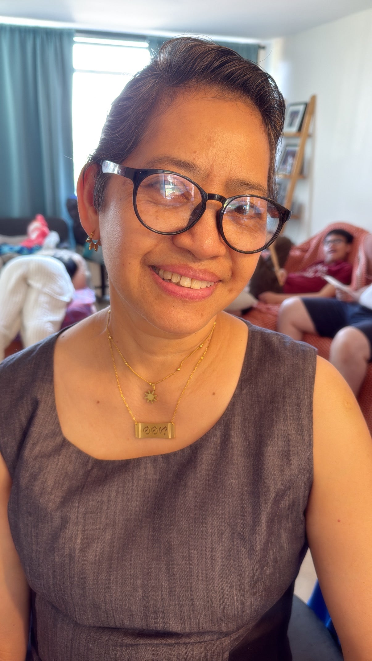 A candid lifestyle photo of a smiling woman with glasses wearing an Aking Sinta gold Nanay bamboo bar necklace layered with a gold sun charm pendant. The horizontal bar piece featuring cutout Baybayin script for mother sits beautifully against her dark gray sleeveless top in a warm, brightly lit home setting