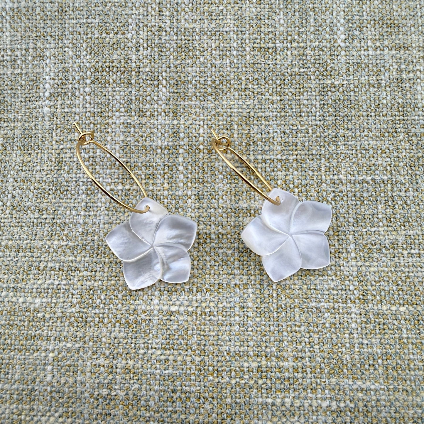 A pair of Aking Sinta gold oval hoop earrings featuring hand-carved Mother of Pearl Kalachuchi (Plumeria) flower drops. The iridescent shell petals are showcased against a neutral woven linen background, highlighting the elegant luster and organic tropical design of the jewelry