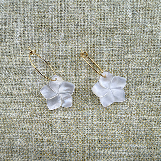 A pair of Aking Sinta gold oval hoop earrings featuring hand-carved Mother of Pearl Kalachuchi (Plumeria) flower drops. The iridescent shell petals are showcased against a neutral woven linen background, highlighting the elegant luster and organic tropical design of the jewelry
