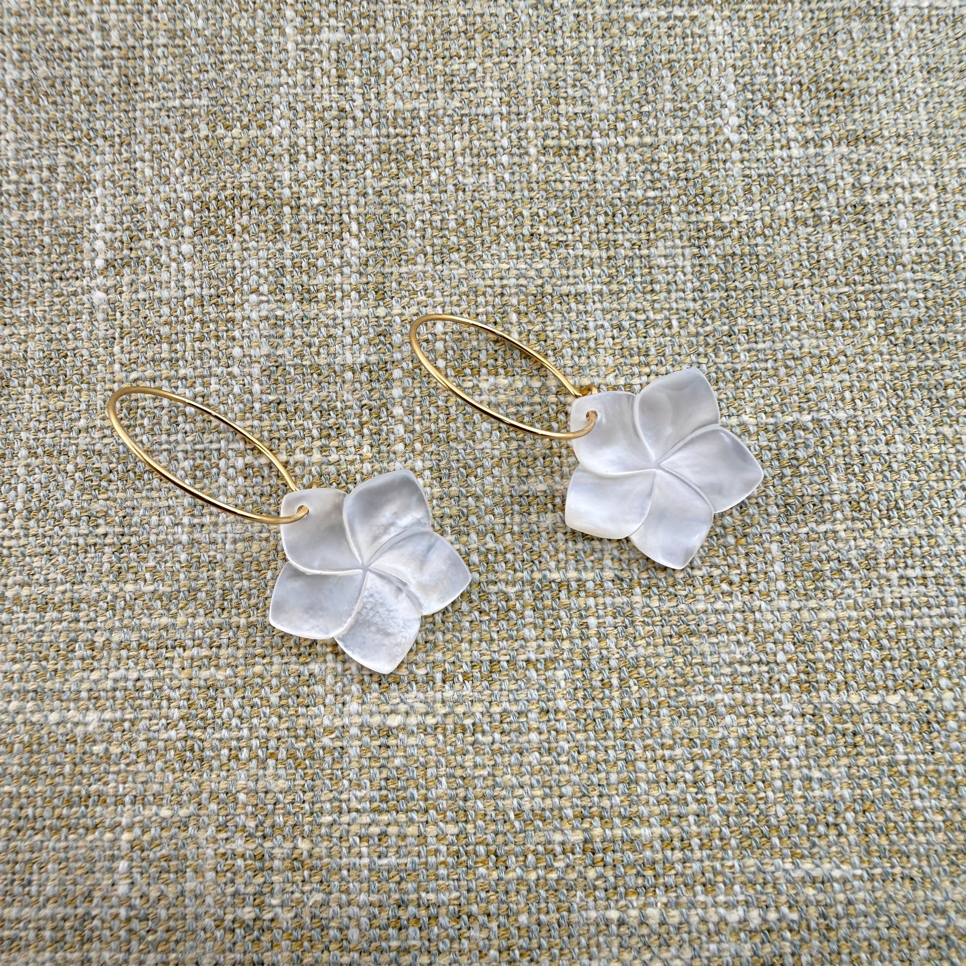 A pair of Aking Sinta gold oval hoop earrings featuring hand-carved Mother of Pearl Kalachuchi (Plumeria) flower drops. The iridescent shell petals are showcased against a neutral woven linen background, highlighting the elegant luster and organic tropical design of the jewelry