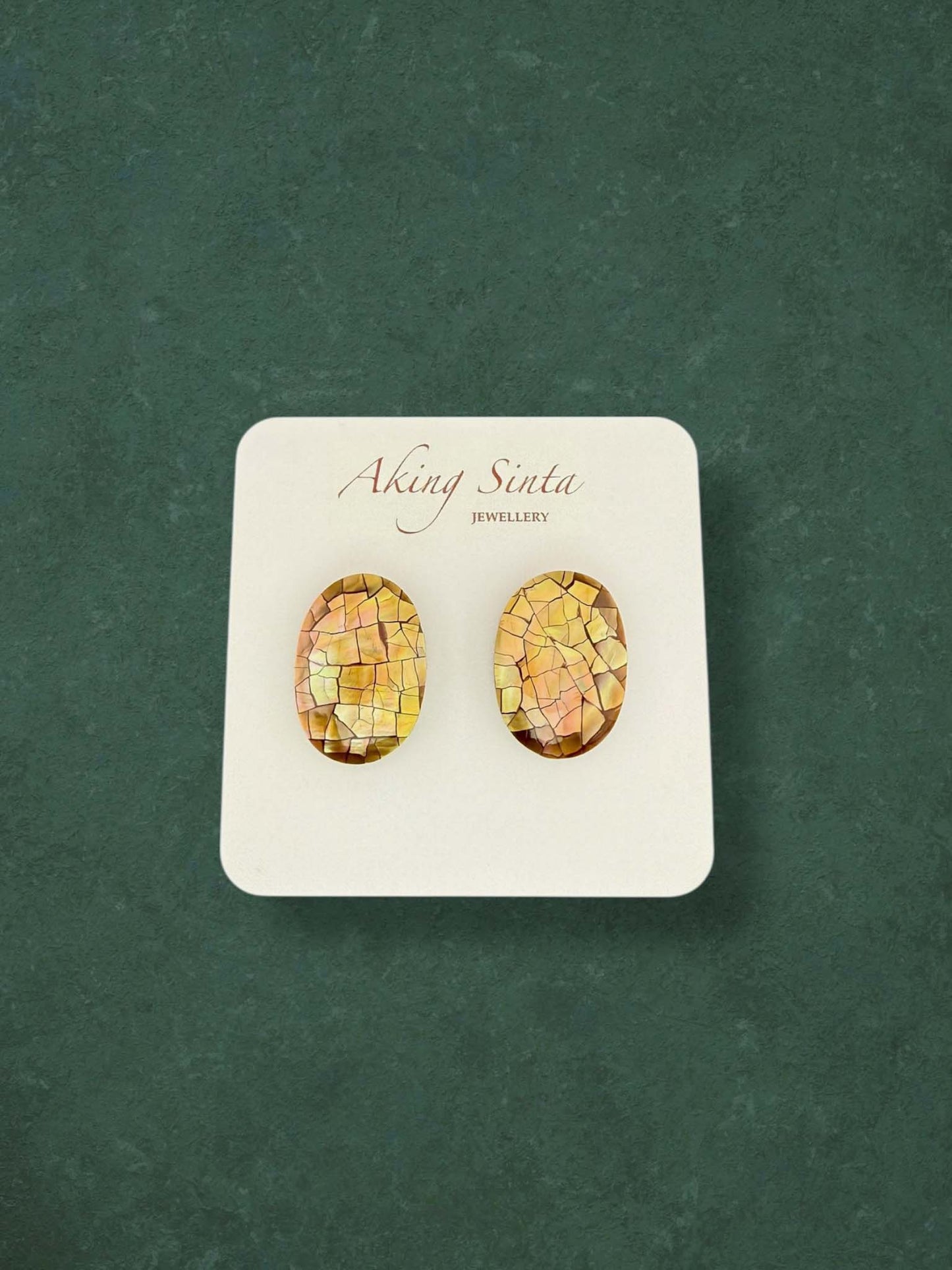 A pair of Aking Sinta oval mosaic mother of pearl stud earrings in warm gold and honey tones. These artisan shell inlay earrings feature a unique cracked mosaic pattern on a white display card