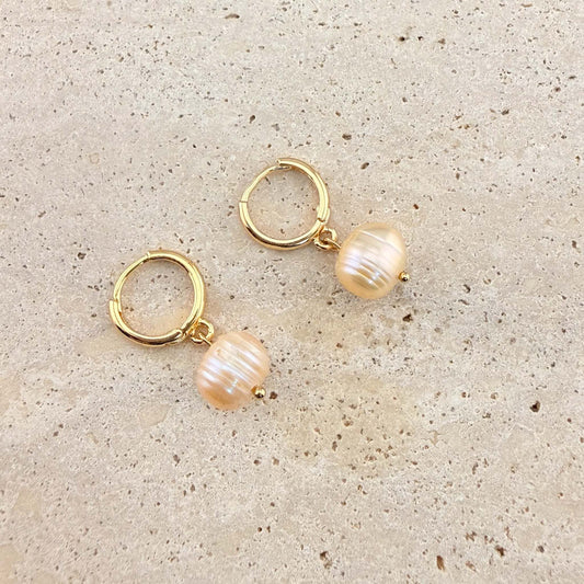 A pair of Aking Sinta gold huggie hoop earrings featuring peach-toned freshwater pearl drops. The round, lustrous pearls hang from polished gold hoops, displayed on a natural light-colored stone surface.