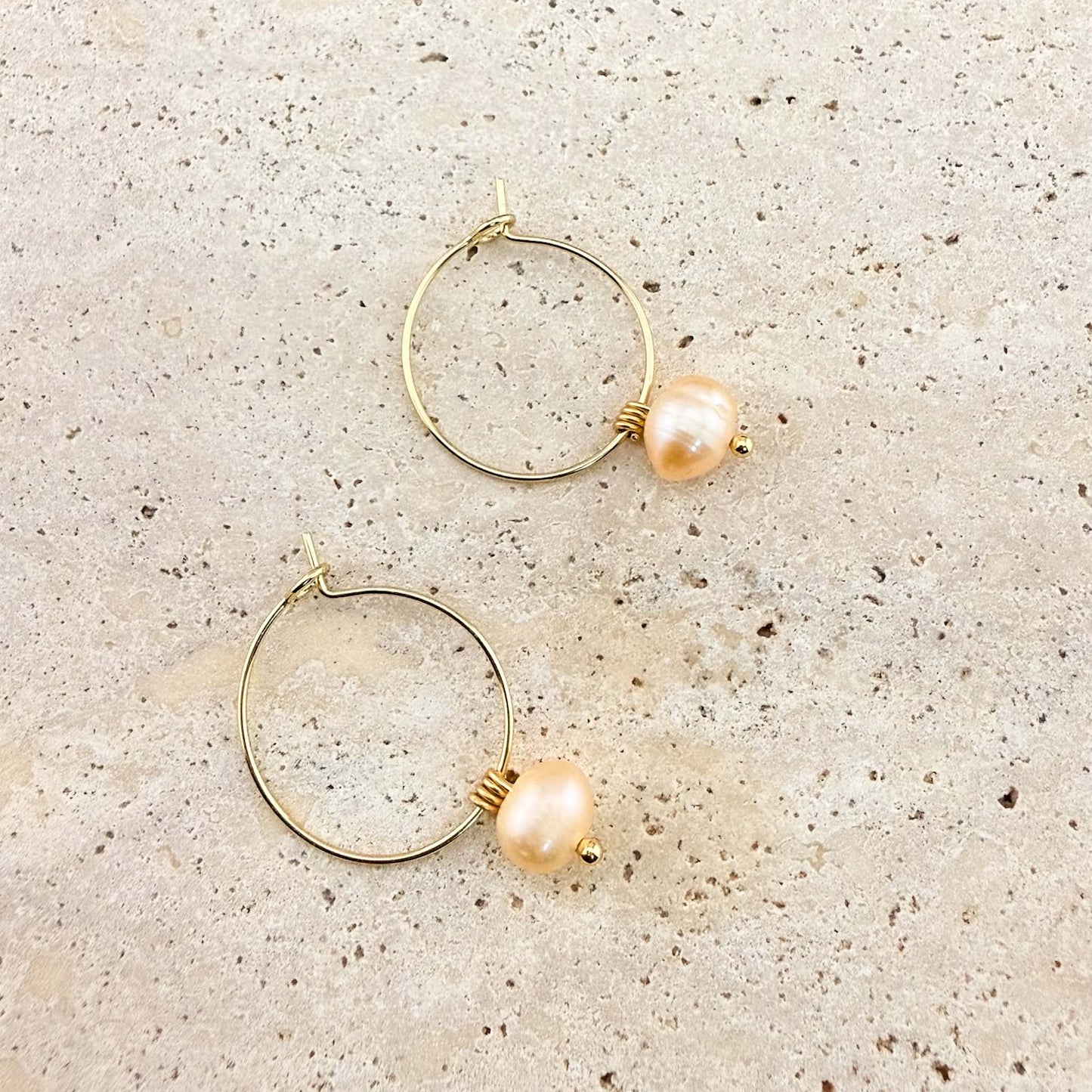 A pair of Aking Sinta gold huggie hoop earrings with peach freshwater pearl drops. One earring is unlatched to show the hinge closure, highlighting the lustrous round pearls on a light-colored natural stone background.