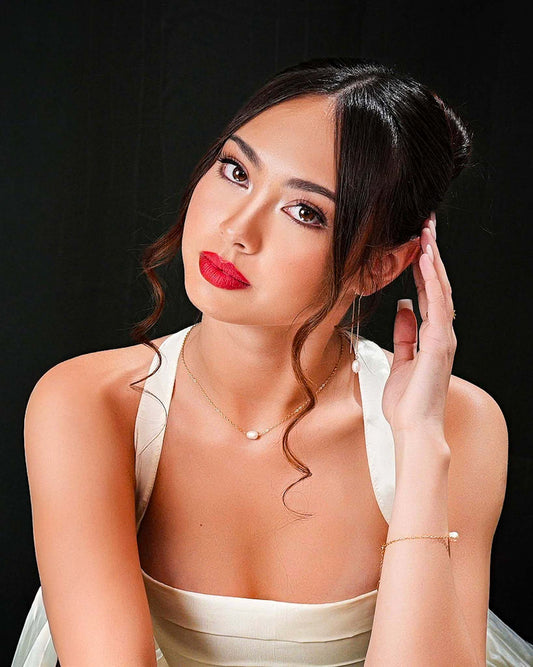 A portrait of a model elegantly wearing a matching Aking Sinta gold jewelry set, featuring a delicate single-pearl necklace, a matching gold chain bracelet with a pearl accent, and gold threader earrings with a single pearl drop. The model is dressed in a white top against a solid black background, highlighting the sophisticated, minimalist beauty of the pearl jewelry collection