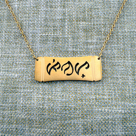 An Aking Sinta gold horizontal bar necklace centered on a textured blue and gold woven linen background. The matte gold pendant features a curved bamboo frame design with the word "Pinay" (Filipina) elegantly cut out in traditional script-like Baybayin characters, suspended from a fine gold link chain.