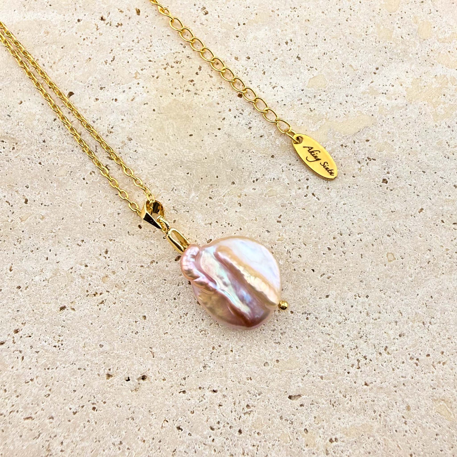 A gold Aking Sinta necklace featuring a luminous, organic-shaped purple keshi pearl pendant. The iridescent pearl displays soft lavender and pink tones, suspended from a polished gold bail and delicate cable chain. The chain includes an adjustable extender finished with a branded "Aking Sinta" oval metal tag, set against a light, textured stone surface.