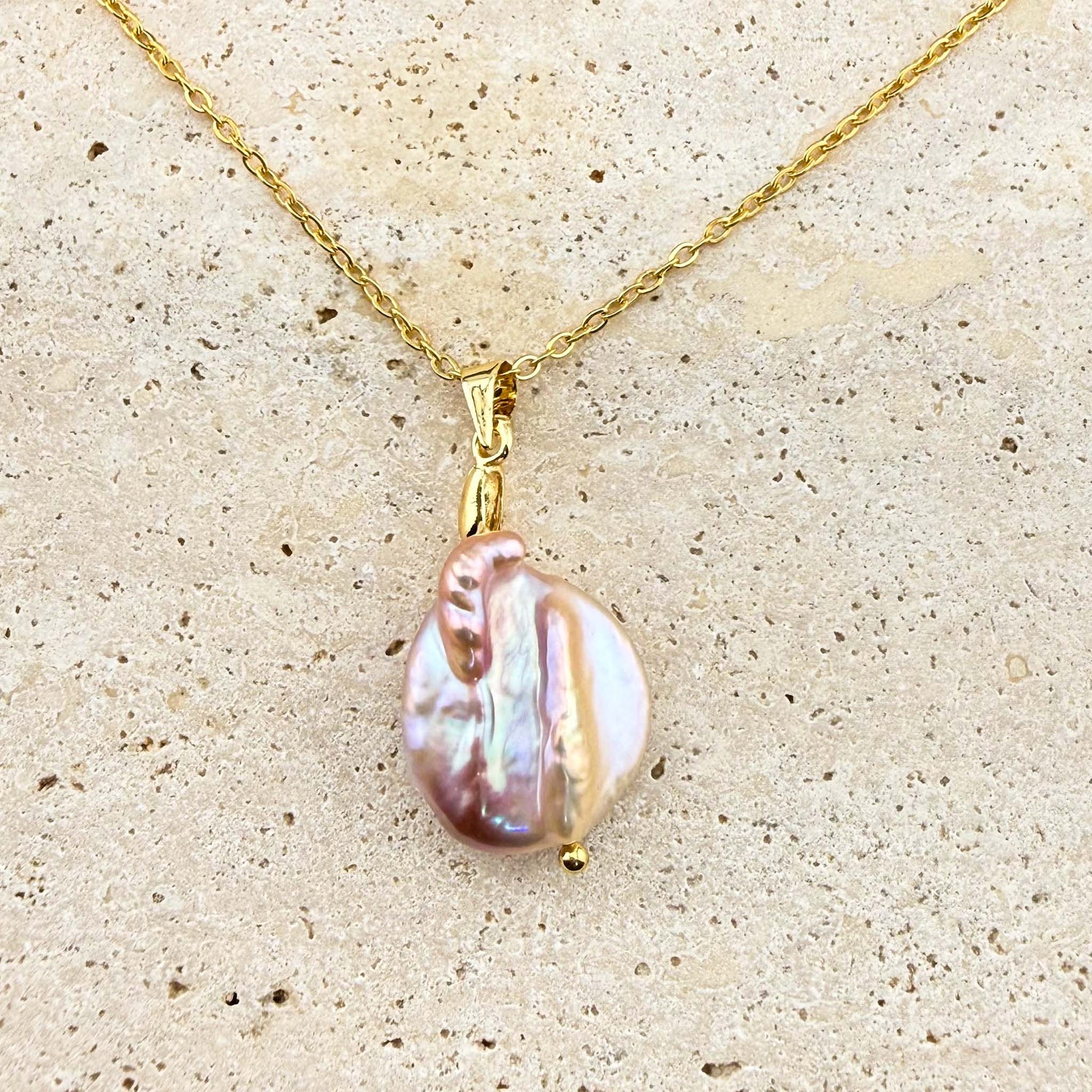 A gold Aking Sinta necklace featuring a luminous, organic-shaped purple keshi pearl pendant. The iridescent pearl exhibits subtle pink and lavender hues, suspended from a polished gold bail and a delicate gold cable chain, displayed on a light, textured stone surface.