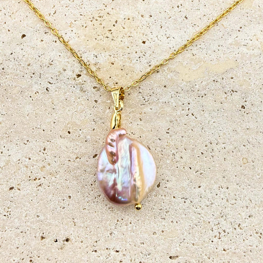 A gold Aking Sinta necklace featuring a luminous, organic-shaped purple keshi pearl pendant. The iridescent pearl exhibits subtle pink and lavender hues, suspended from a polished gold bail and a delicate gold cable chain, displayed on a light, textured stone surface.