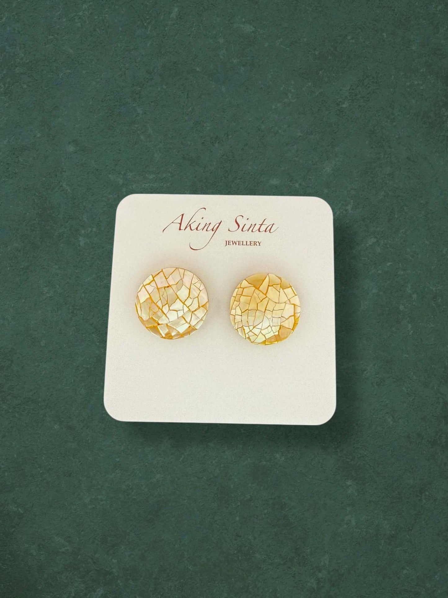 A pair of Aking Sinta round mosaic mother of pearl stud earrings in a warm golden yellow hue. These circular shell inlay earrings feature a textured cracked-stone pattern on a white branded display card