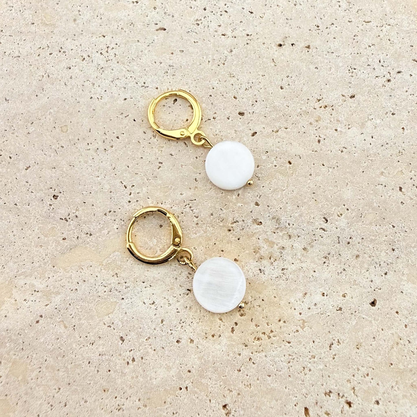 A pair of Aking Sinta gold huggie hoop earrings featuring flat, circular mother of pearl disc charms. The iridescent white shell drops are displayed on a natural, sand-colored stone surface.
