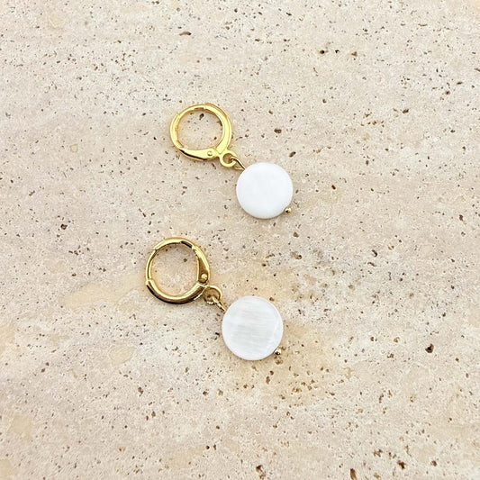 A pair of Aking Sinta gold huggie hoop earrings featuring flat, circular mother of pearl disc charms. The iridescent white shell drops are displayed on a natural, sand-colored stone surface.