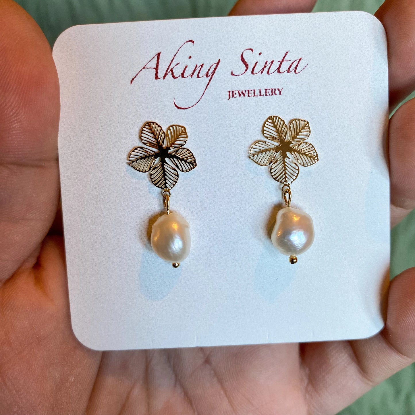 A pair of gold-tone Aking Sinta statement earrings presented on a branded white display card. Each earring features an intricate filigree Sampaguita flower stud with a dangling, organic white baroque pearl drop, held in a hand against a soft green background