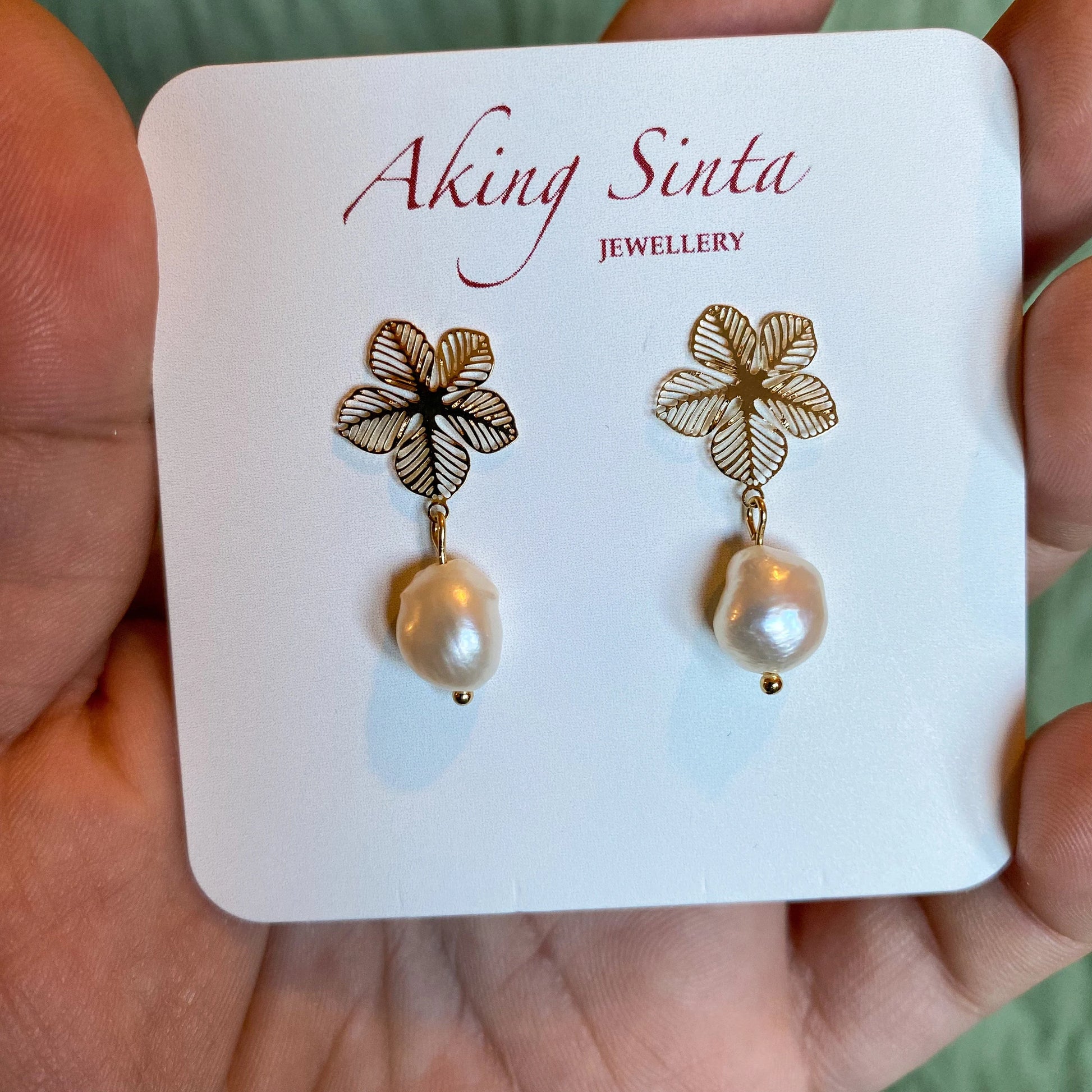 A pair of gold-tone Aking Sinta statement earrings presented on a branded white display card. Each earring features an intricate filigree Sampaguita flower stud with a dangling, organic white baroque pearl drop, held in a hand against a soft green background