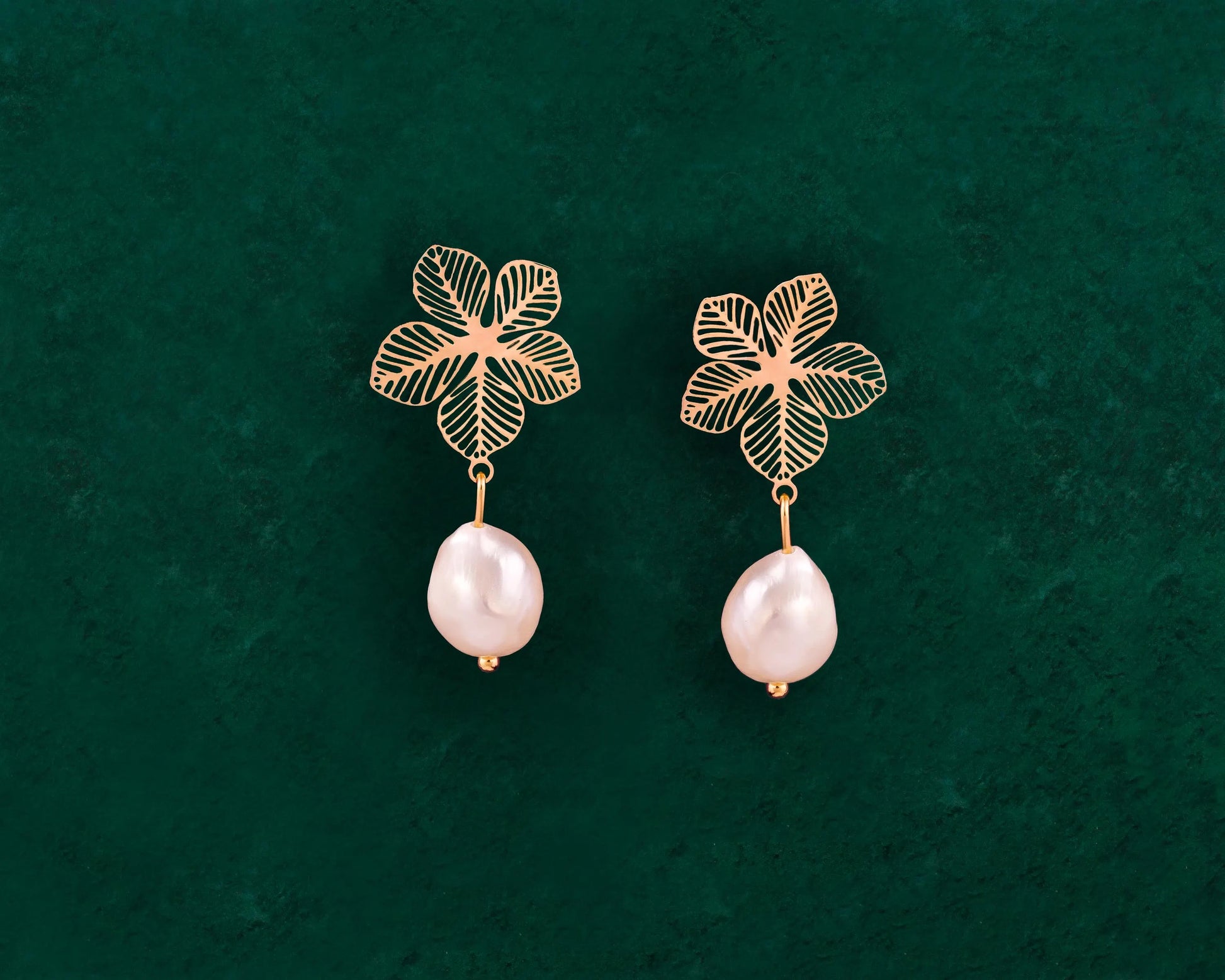 A pair of Aking Sinta statement earrings featuring intricate gold filigree Sampaguita flower studs. Suspended from each flower is a lustrous, organic-shaped white baroque pearl drop. The earrings are presented against a rich, dark green velvet background, highlighting the contrast between the polished gold and the natural pearl sheen