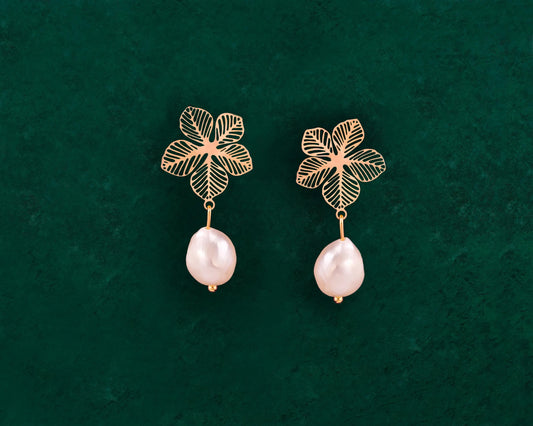 A pair of Aking Sinta statement earrings featuring intricate gold filigree Sampaguita flower studs. Suspended from each flower is a lustrous, organic-shaped white baroque pearl drop. The earrings are presented against a rich, dark green velvet background, highlighting the contrast between the polished gold and the natural pearl sheen