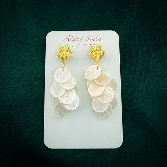 A pair of Aking Sinta statement earrings featuring gold Sampaguita flower studs with intricate petal detailing. Hanging from the studs are cascading clusters of iridescent white Capiz shell discs. The earrings are presented on a white branded display card against a dark green velvet background.