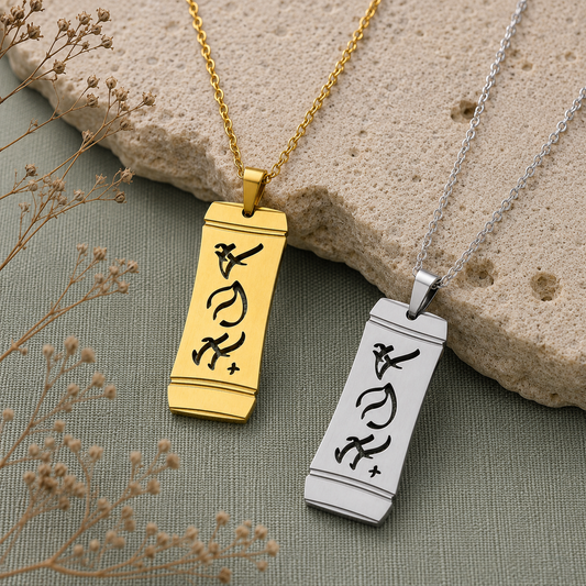 A product shot featuring two Aking Sinta vertical bamboo-style rectangular pendants in gold and silver finishes, displayed side by side against a textured stone slab and sage green fabric backdrop with dried baby's breath. Both pendants showcase the word "Anak" (Child) intricately cut out in stylized traditional Baybayin script characters.
