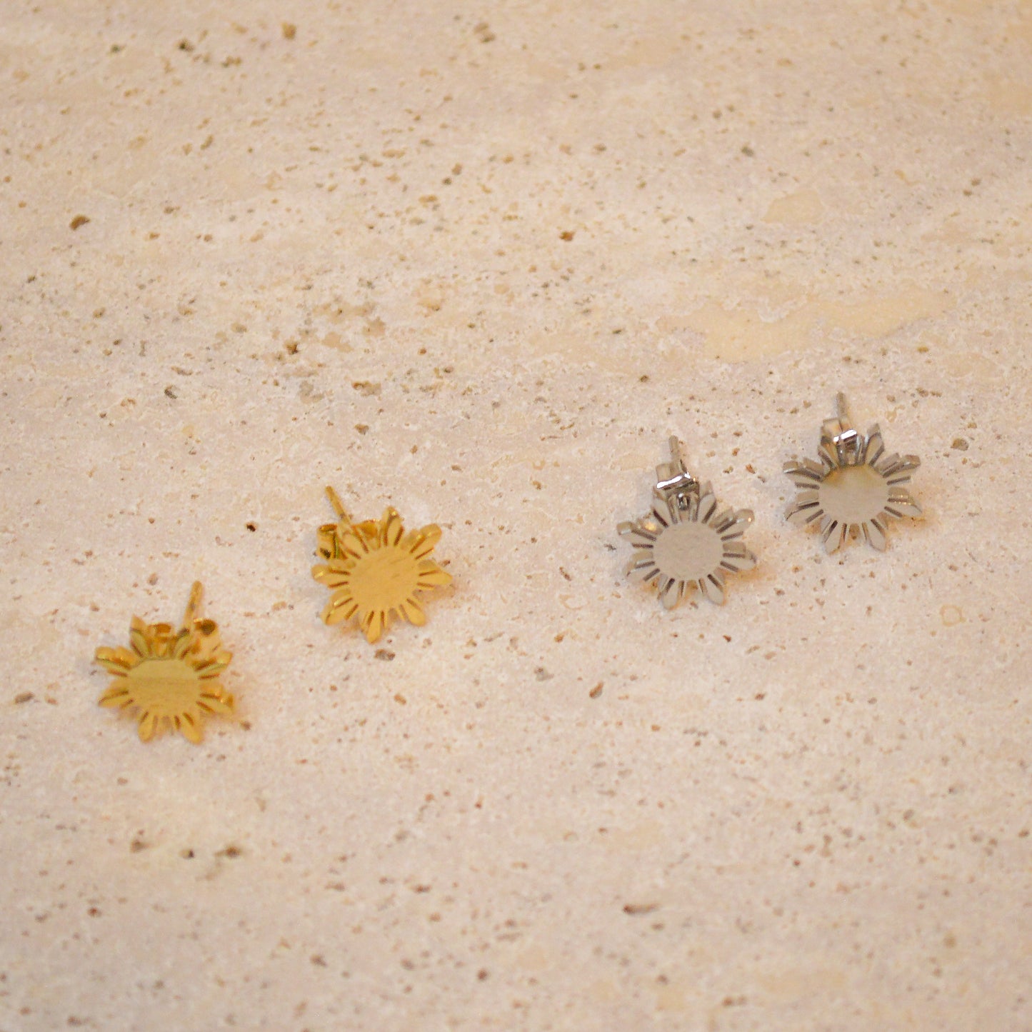 Two pairs of Aking Sinta Araw stud earrings—one in polished gold and one in sleek silver—displayed on a light-colored, textured natural stone surface. The minimalist eight-rayed sun design, inspired by Filipino heritage, is shown from a high-angle perspective to highlight the clean geometric lines and metallic finish of both color options.