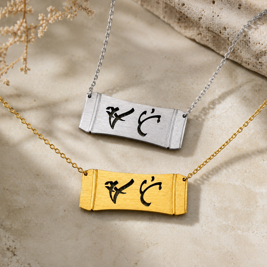 A product display featuring two Aking Sinta horizontal bamboo bar necklaces in matte silver and gold finishes. The parallel curved pendants feature the word "Ate" (Elder Sister) in cutout script-like Baybayin characters, photographed on a light stone surface with subtle shadows and a sprig of dried florals.