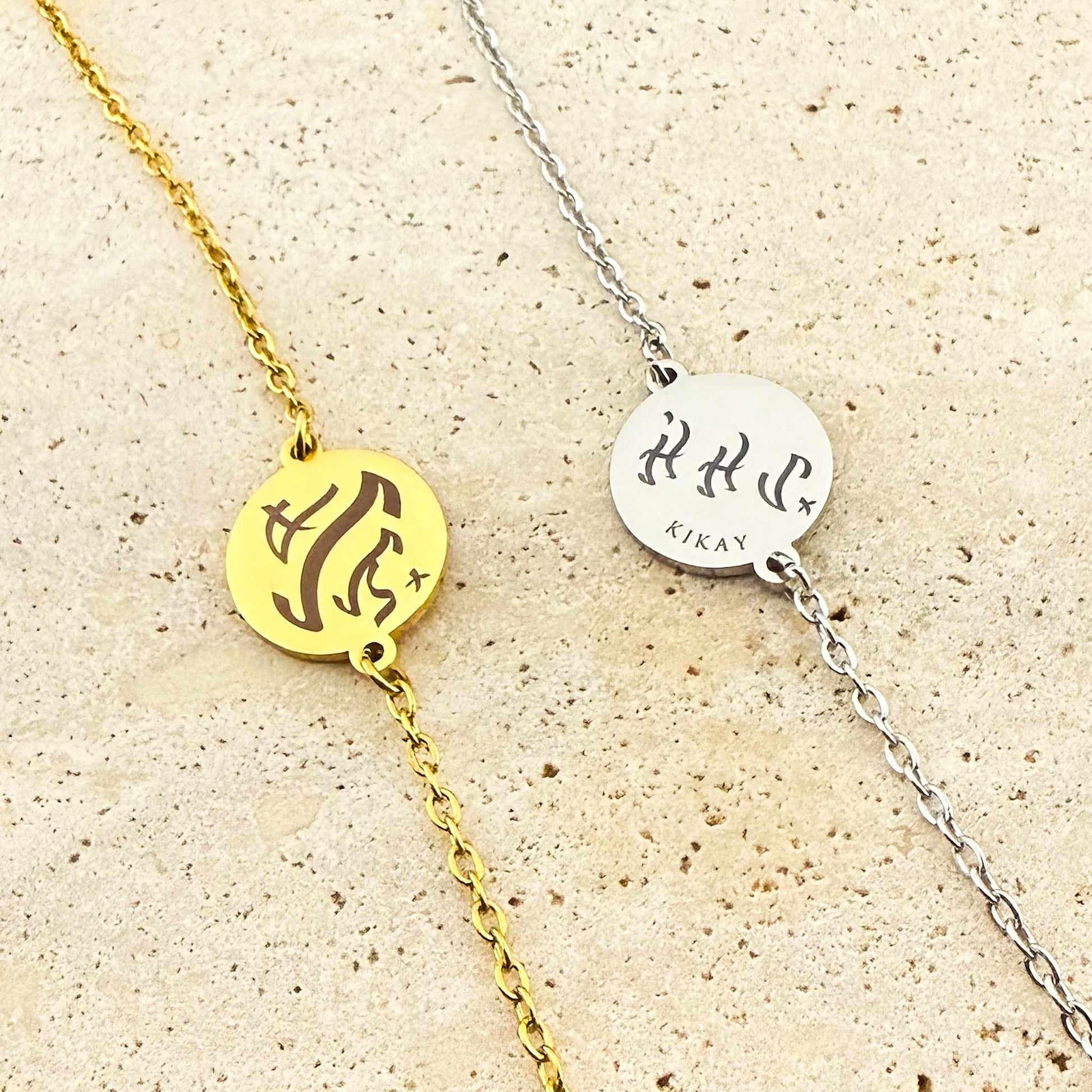 Two Aking Sinta coin bracelets, one in gold and one in silver, featuring bespoke engravings in ancient Filipino Baybayin script. The silver coin includes the word "Kikay" below the script, shown on a light-colored natural stone background
