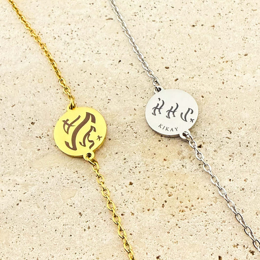 Two Aking Sinta coin bracelets, one in gold and one in silver, featuring bespoke engravings in ancient Filipino Baybayin script. The silver coin includes the word "Kikay" below the script, shown on a light-colored natural stone background