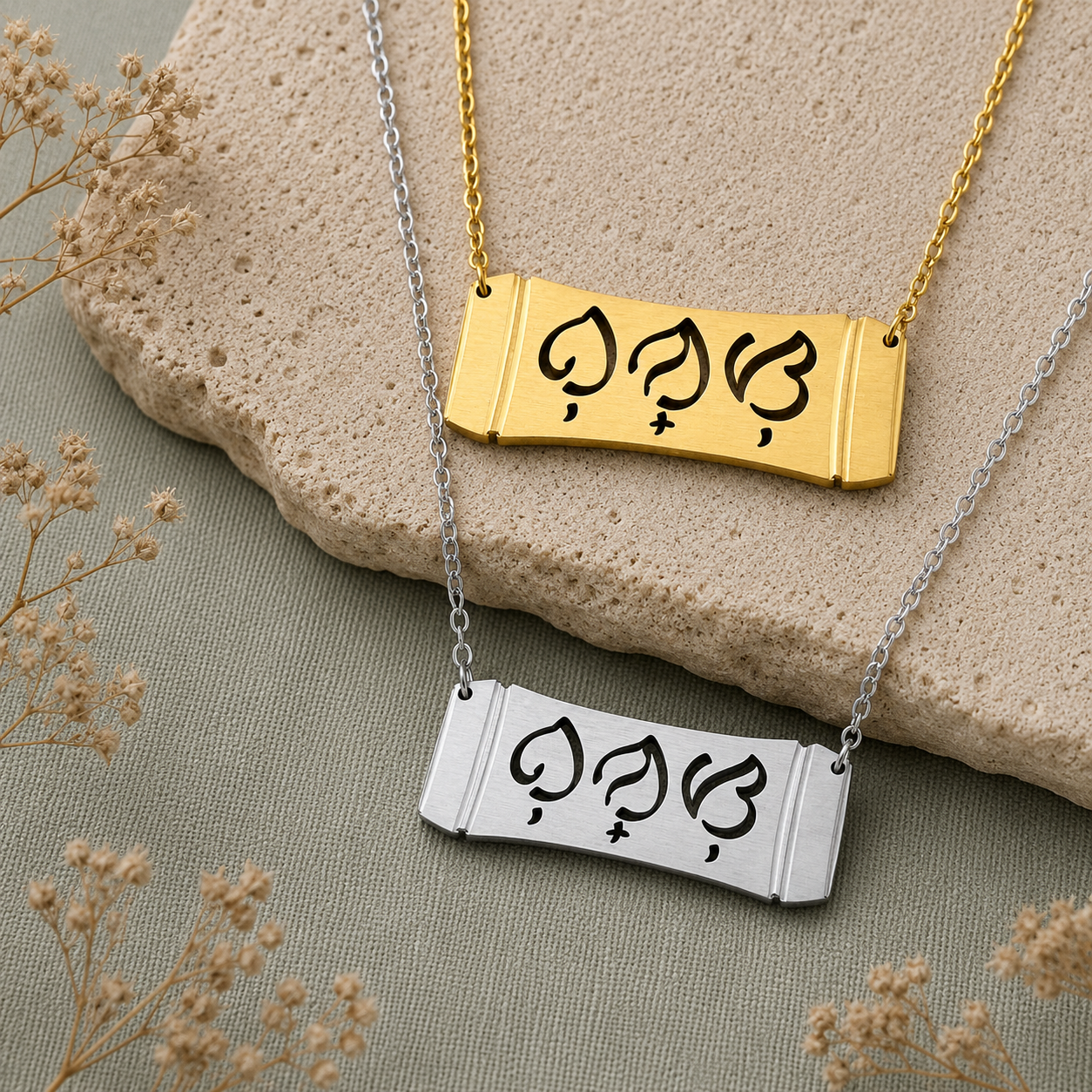 A flat lay product photograph featuring two Aking Sinta horizontal bamboo bar necklaces in matte gold and silver finishes. The parallel curved pendants display the word "Bunso" (Youngest Child) in clean cutout Baybayin script characters, arranged over a light beige textured stone slab and sage green fabric backdrop adorned with dried baby's breath.