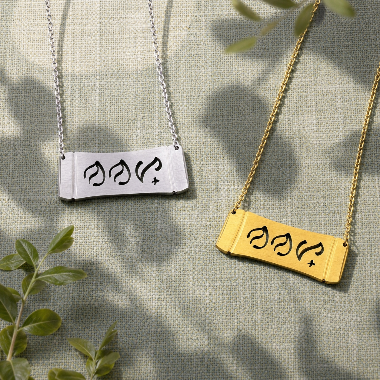 showcasing two Aking Sinta horizontal bamboo bar necklaces in matte silver and gold finishes. The parallel curved pendants feature the word "Nanay" (Mother) delicately cut out in traditional script-like Baybayin characters, arranged on a textured sage green fabric surface beautifully accented by soft leaf shadows and green sprigs.