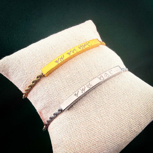 A pair of Aking Sinta twisted rope chain bracelets in gold and silver finishes. Each bracelet features a curved metal bar custom-engraved with ancient Filipino Baybayin script, displayed together on a small cream-colored jewelry pillow