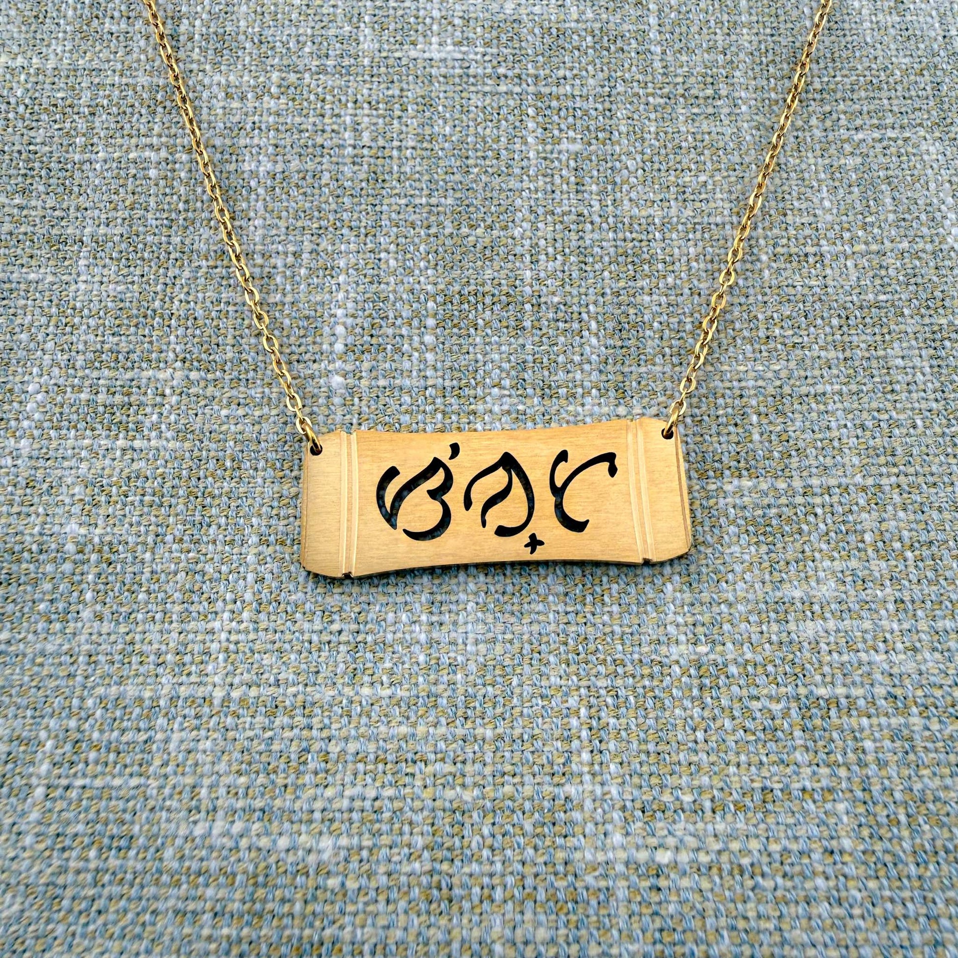 An Aking Sinta gold horizontal bar necklace arranged on a textured blue and gold woven linen background. The matte gold pendant features a curved bamboo frame silhouette with the brand's namesake word "Sinta" (Beloved) beautifully cut out in traditional script-like Baybayin characters, attached to a fine gold link chain.