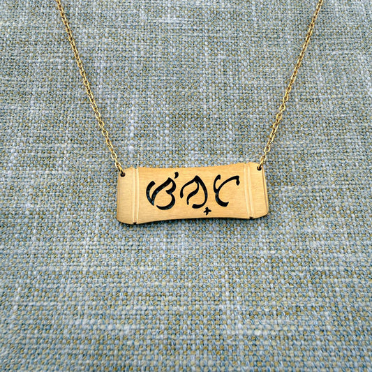 An Aking Sinta gold horizontal bar necklace arranged on a textured blue and gold woven linen background. The matte gold pendant features a curved bamboo frame silhouette with the brand's namesake word "Sinta" (Beloved) beautifully cut out in traditional script-like Baybayin characters, attached to a fine gold link chain.
